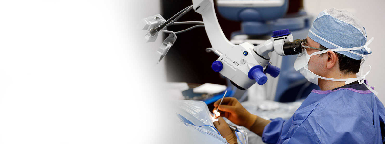 Surgeon in blue scrubs and a cap uses a surgical microscope in an operating room.