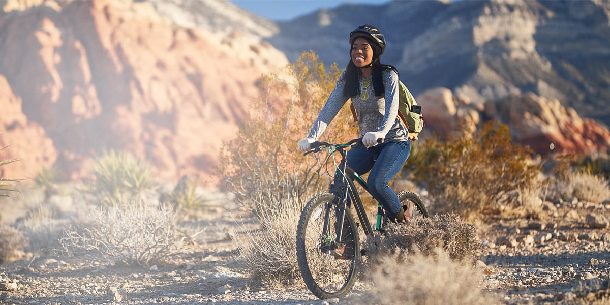A woman riding a mountain bike through a desert landscape, wearing a helmet and smiling with sharp, comfortable vision, surrounded by red rock formations and desert plants.