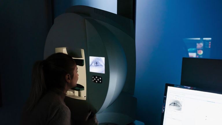 A person sits beside a large MRI scanner in a dim room, with a computer monitor on a desk to the right.