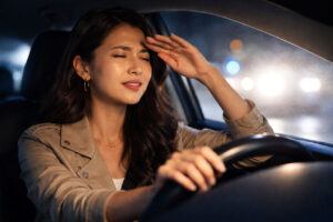 A woman sits in the driver's seat at night with her hand on her forehead, appearing stressed or tired.